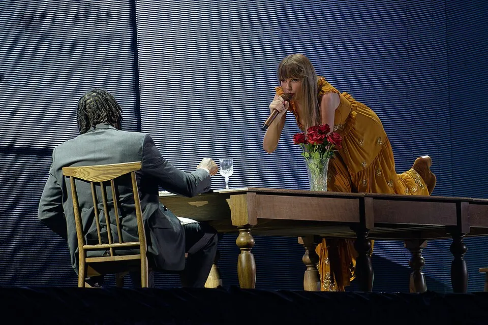 A woman in a mustard dress singing across a dinner table on a stage.