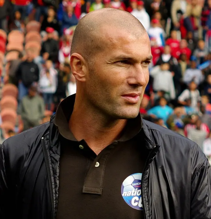 A bald man in a dark jacket at a football stadium.