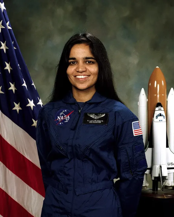 A smiling woman in a blue flight suit beside a flag and a shuttle model.