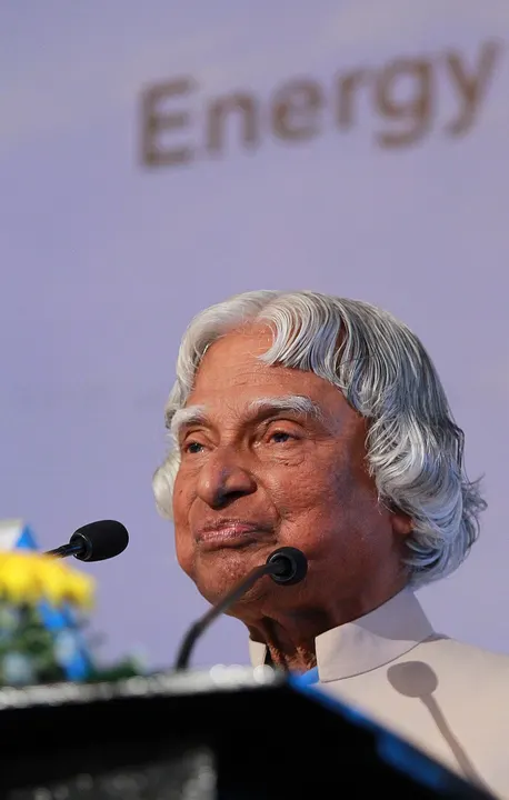 A smiling elderly man with swept-back grey hair speaking at a podium.