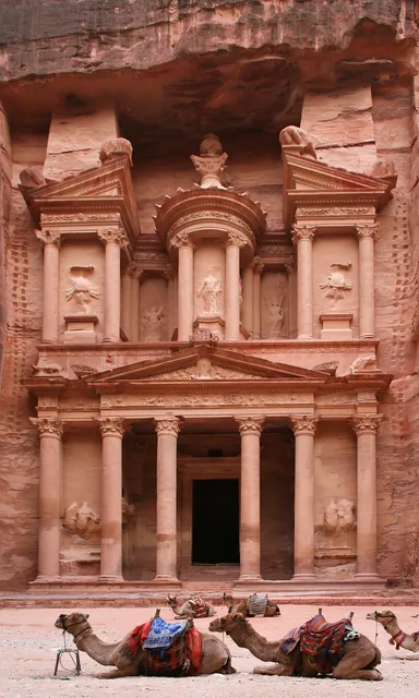 Ornate columned facade carved into a red rock cliff, with camels resting in front
