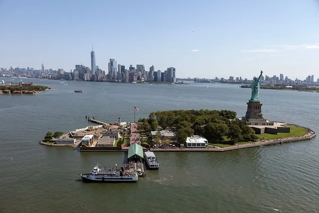 Aerial view of a small island with a torch-bearing statue, a city skyline across the water