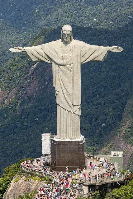 Huge statue of a robed figure with outstretched arms on a mountain summit, visitors on the platform below
