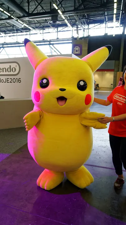 A giant fluffy yellow costume figure with long black-tipped ears and red cheeks at a game convention booth.