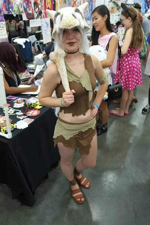 A cosplayer in a bone-white animal-skull helmet and a brown leaf-like top and skirt, holding a long bone over one shoulder.