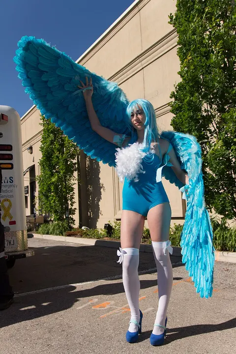 A cosplayer in a bright blue leotard with huge feathered blue wings and white stockings, standing in a street.