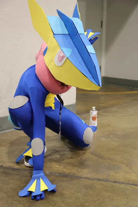 A cosplayer crouching in a blue bodysuit with an angular yellow and blue head, a pink scarf-like tongue and webbed hands.