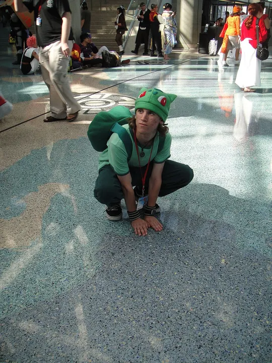 A crouching cosplayer in a green hat with red eyes and a big green backpack shaped like a shell, on a polished floor.