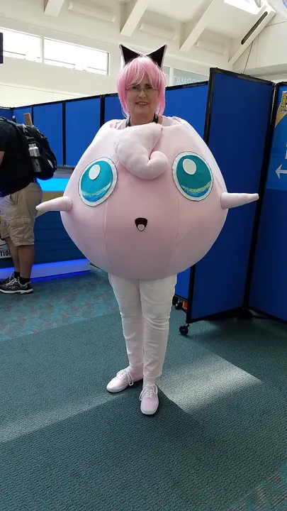 A smiling cosplayer in a huge round pink costume with large teal eyes, pointed side ears and a curled tuft, with pink hair and cat ears.