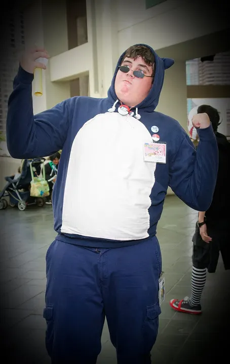 A cosplayer in a dark blue hoodie with a cream belly patch and pointed ears, wearing sunglasses and flexing an arm.
