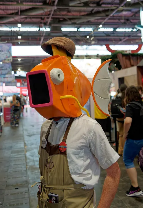 A person in a giant orange fish-head costume with a wide pink mouth and a grey fin, wearing a flat cap and a shop apron at a busy event.