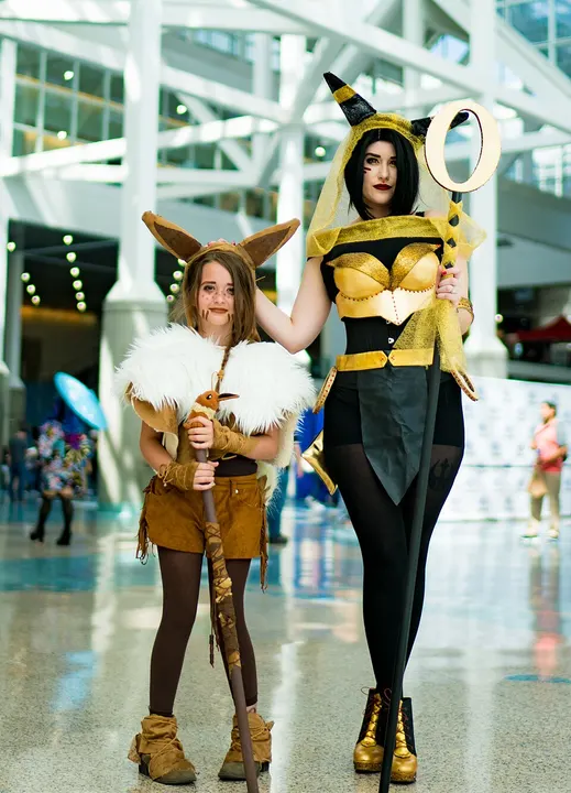Two cosplayers at a convention: a child in a brown outfit with a fluffy cream collar and fox-like ears on the left, and a taller woman in black and gold with a ring-topped staff.