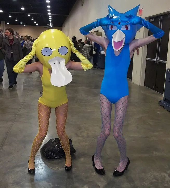 Two cosplayers in latex bodysuits and bird-like beak masks, a yellow one on the left and a blue one on the right, in a convention hall.