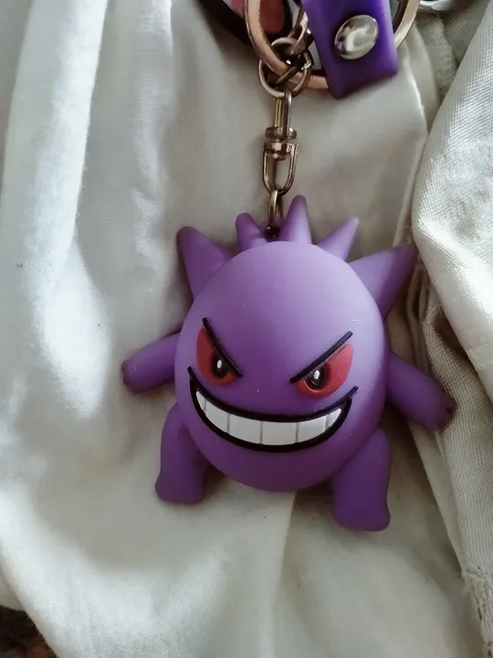 A purple rubbery keychain figure with spikes, red eyes and a wide toothy grin hanging on a cream cloth.