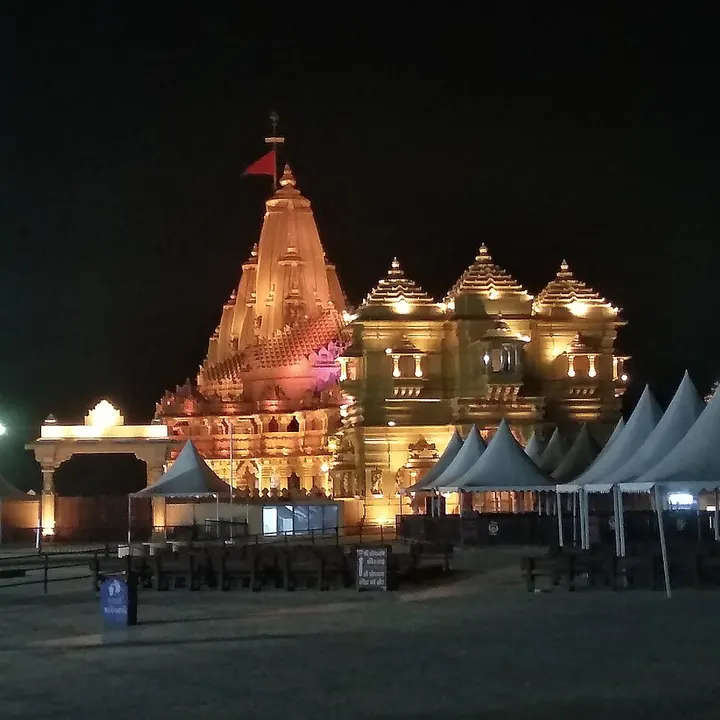 A tall golden-lit temple with a red flag on its spire stands at night, with white tents in the open ground in front.