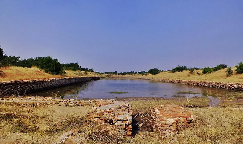 A long, brick-lined rectangular basin holding some water lies in dry grassland under a blue sky.