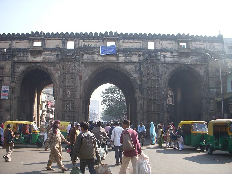 A huge old triple-arched stone gateway towers over a busy street with auto-rickshaws and pedestrians.