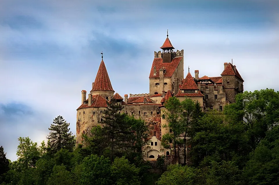 A hilltop castle with orange-tiled towers and turrets among green trees under a moody sky.