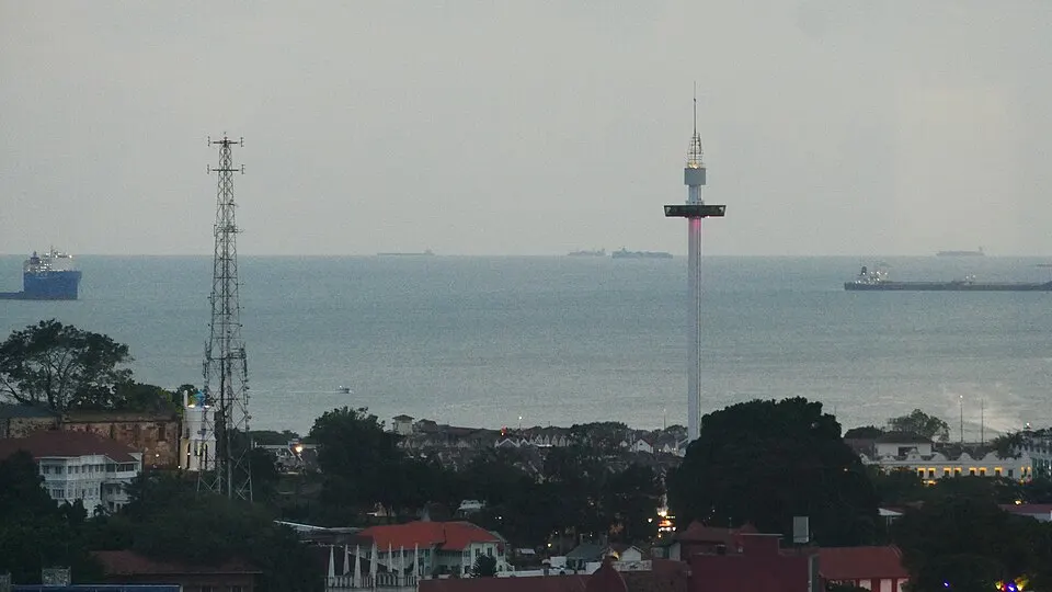 Cargo ships on a grey sea seen from a coast, with a tall slim tower and rooftops in the foreground.