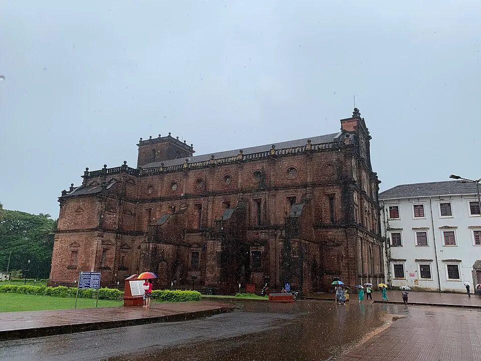 A large dark red stone church with a tiled roof on a rainy day, with people holding umbrellas.