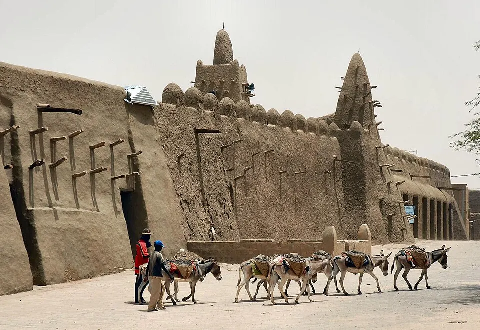 A line of donkeys carrying loads along a street beside a tall wall of mud with wooden beams sticking out.