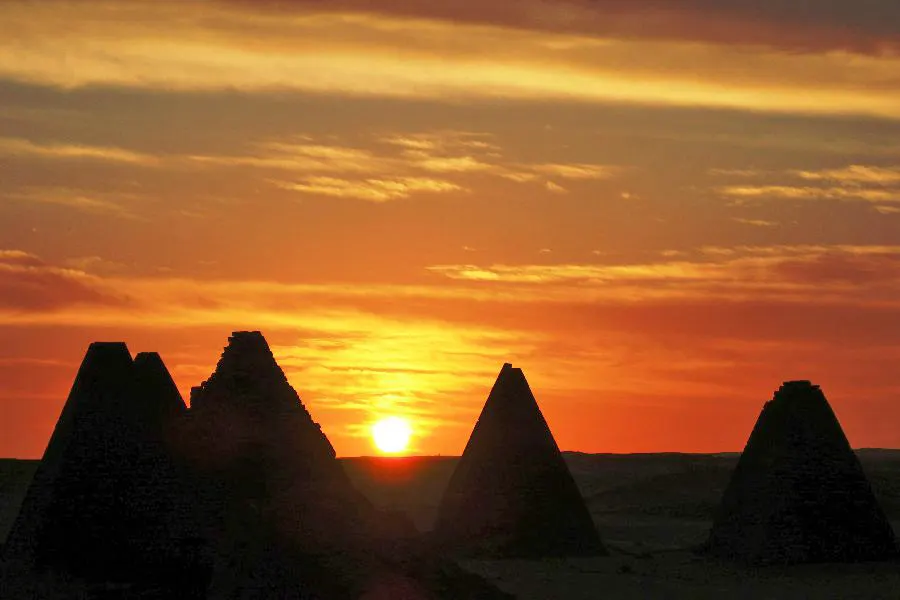 The dark silhouettes of several steep-sided pyramids against an orange sunset.