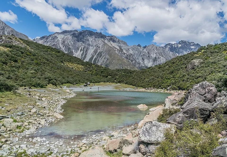 A turquoise mountain lake with stony shores below rugged grey peaks and green slopes.