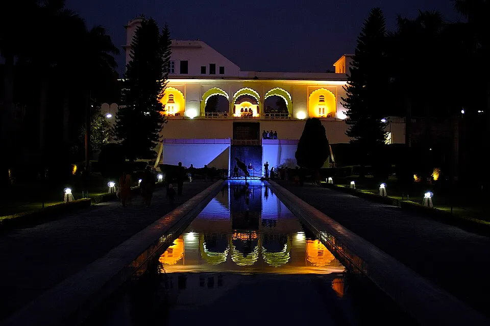 A lit-up palace pavilion with yellow arches is mirrored in a long channel of water between dark trees at night.