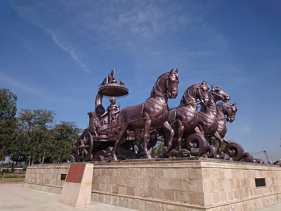 A large bronze sculpture of a chariot pulled by four galloping horses, with a figure standing under a canopy, stands on a stone platform.