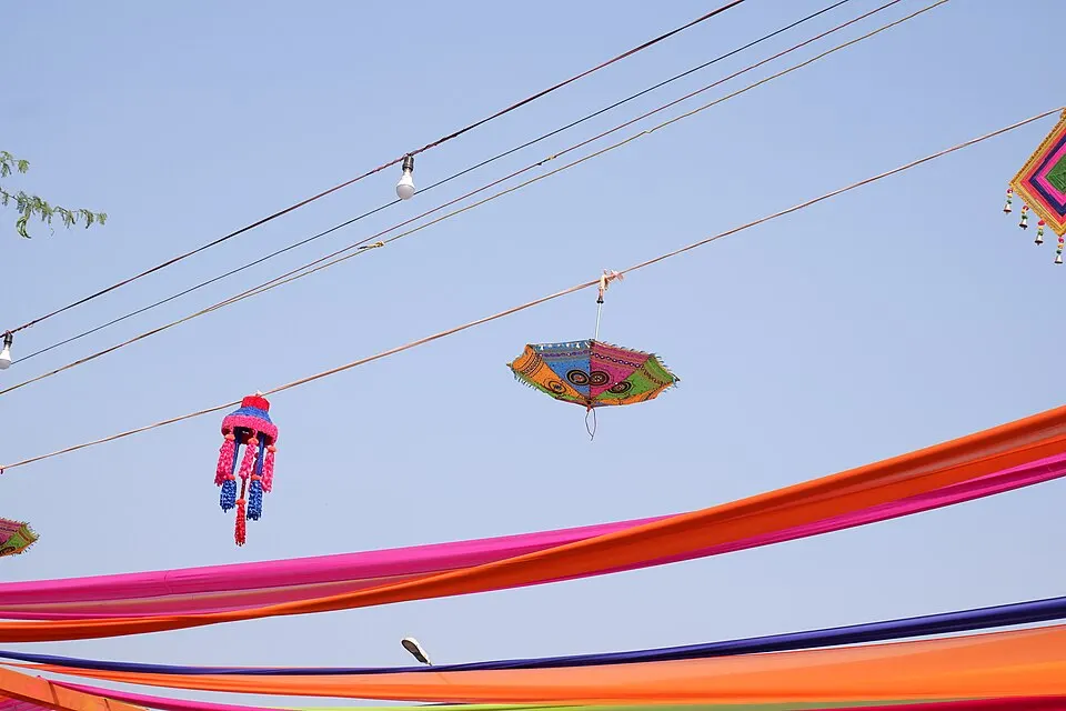 Colourful embroidered umbrellas, tasselled lanterns and bright fabric streamers hang from wires against a pale blue sky.