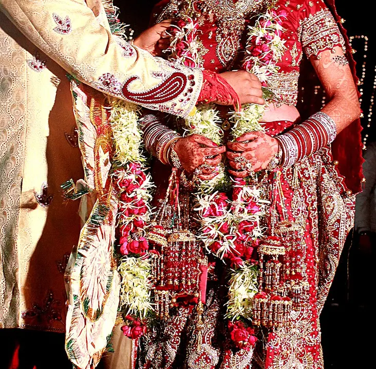 A bride in a red embroidered lehenga and a groom in a cream sherwani stand close together wearing flower garlands, hands joined.