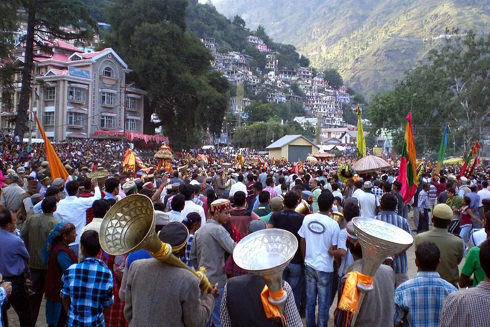 A dense crowd with long brass horns and tall colourful flags fills a street below a steep hillside of houses.