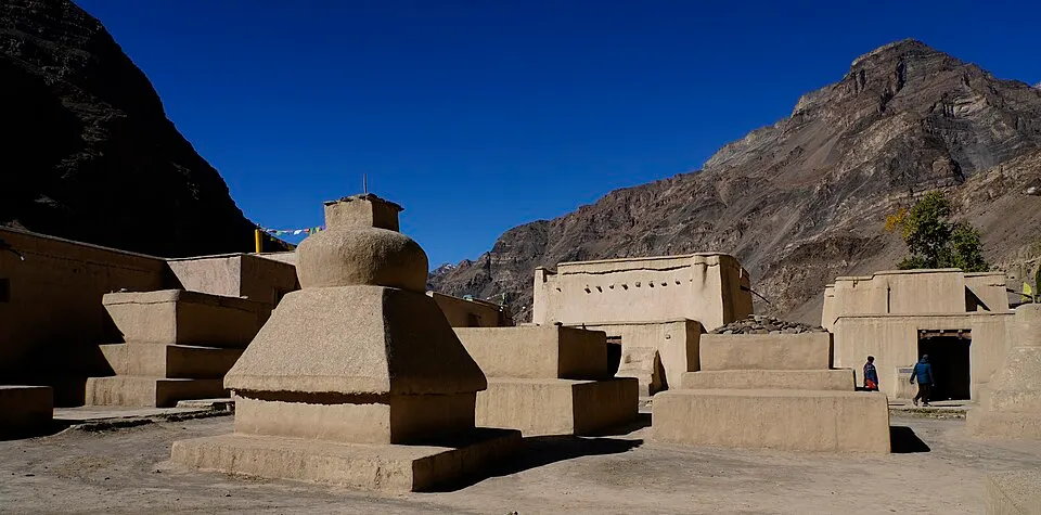 Mud-coloured stupas and low flat-roofed buildings sit in a barren mountain valley under a deep blue sky.