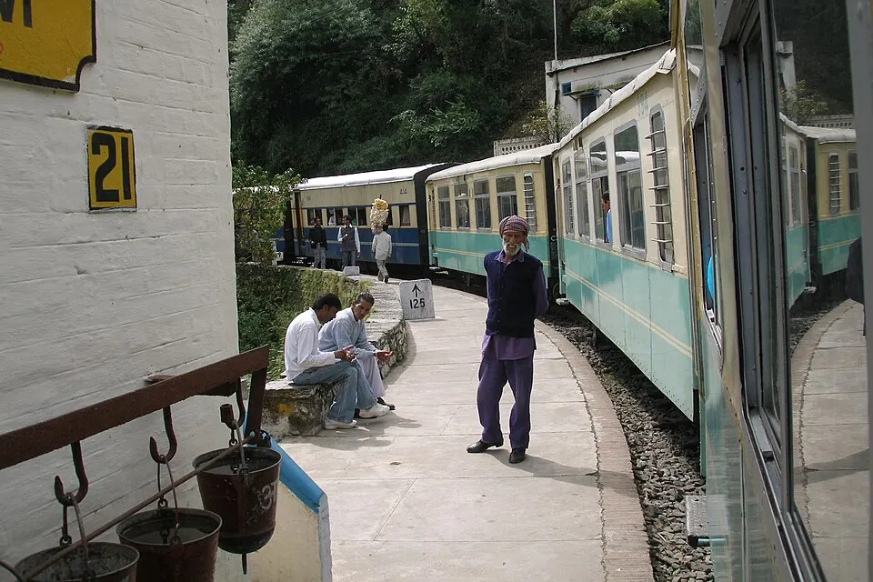 A narrow-gauge train in teal and cream curves past a small hillside platform where a few men stand and sit.