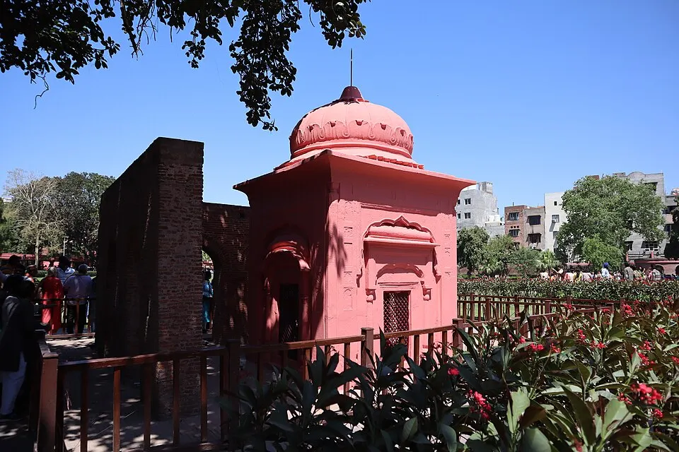एक बाग में गुलाबी रंग का गुंबदवाला छोटा स्मारक, उसके पास ईंट की पुरानी दीवार और आसपास लोग।