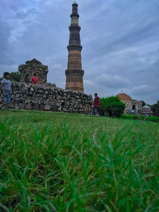बादलों भरे आसमान के नीचे घास के मैदान के पीछे खड़ी लाल-भूरे पत्थर की ऊँची मीनार।