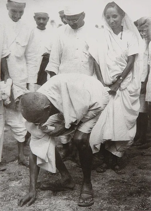 A black-and-white photo of a bald man in white bending to pick something up from the ground as others look on.