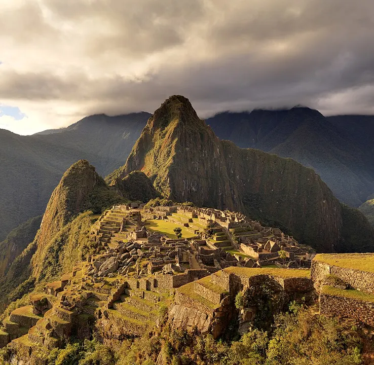 Ancient stone terraces and ruins on a green ridge beneath a steep pointed mountain and stormy clouds.
