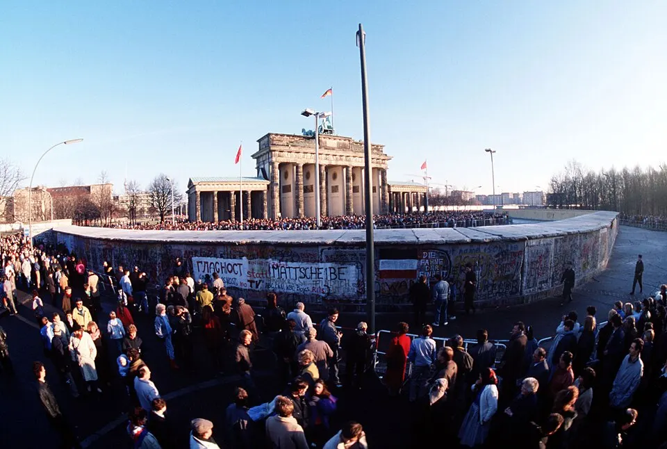 A big crowd gathers by a graffiti-covered concrete wall, with a columned gate behind it.