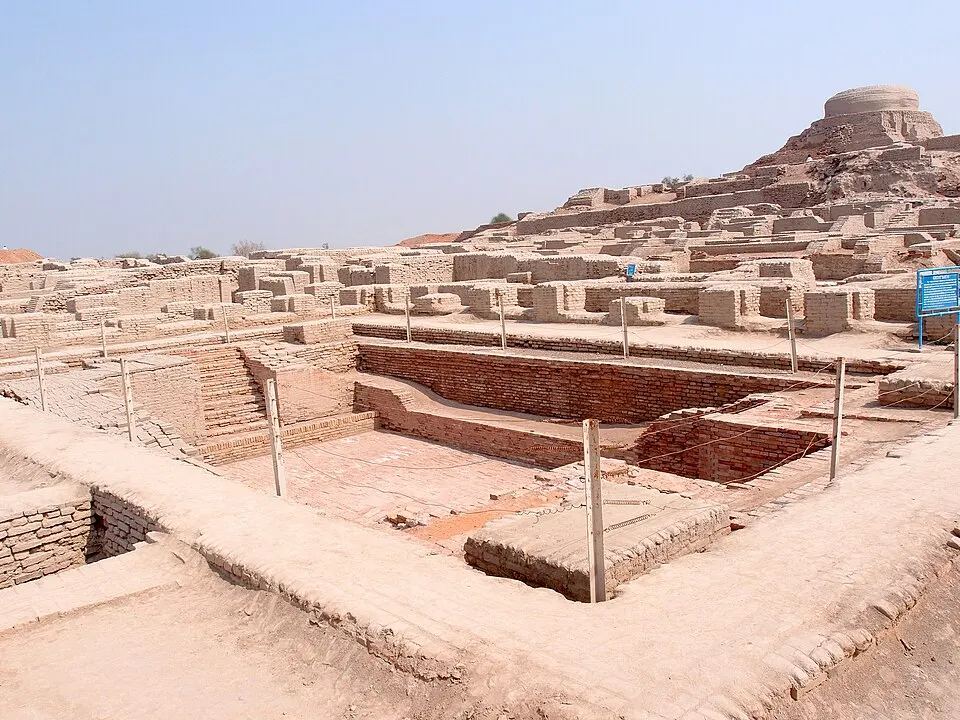 Excavated ancient brick ruins with a large sunken brick pool in the foreground and a mound topped by a round structure.