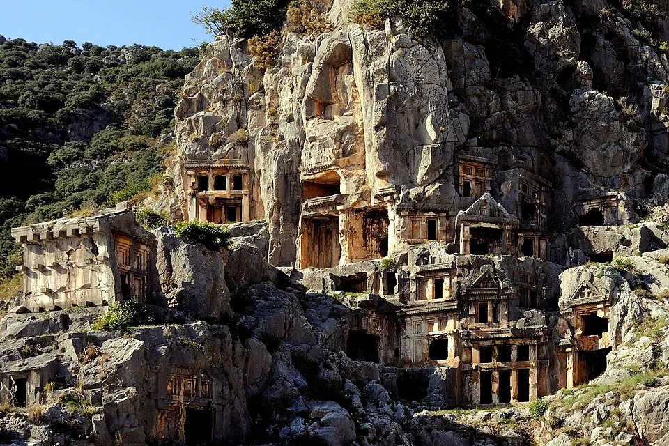 Ancient tombs carved into a steep cliff face with temple-style fronts, beside green hills.