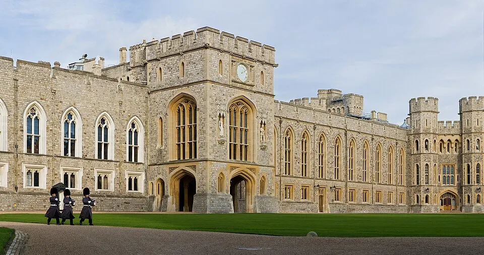 A large castle courtyard with crenellated towers, a green lawn and three guards in dark coats walking past.
