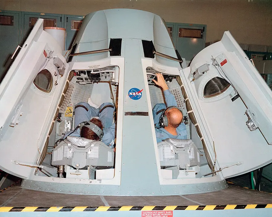 Two men in blue flight suits lying inside an open mock spacecraft capsule with its two hatches swung wide.