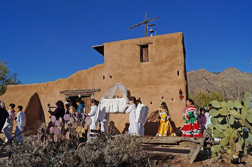 Children in white angel costumes and colourful dresses carrying a small shrine in procession past an adobe church with cactus.