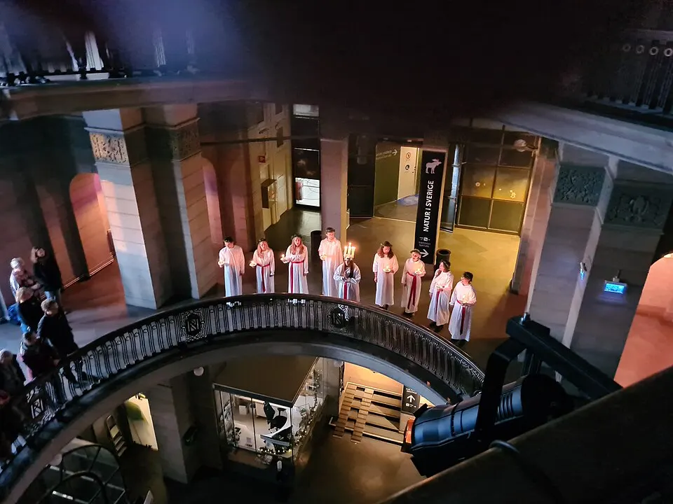 Young people in white robes with red sashes and candles, one wearing a crown of candles, standing in a museum hall.