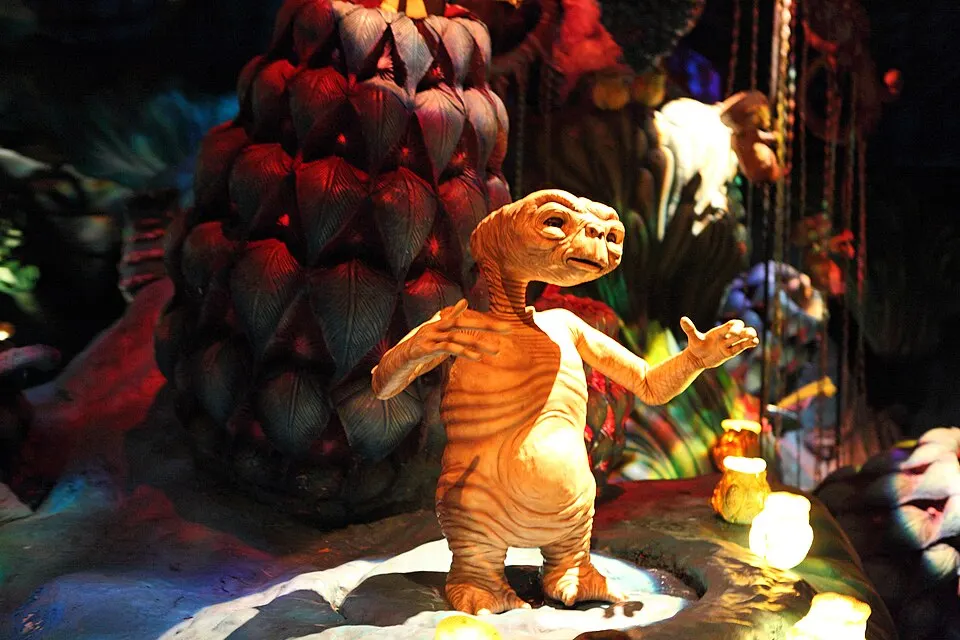 A small brown-skinned alien figure with big eyes and long thin arms stands among glowing lamps and giant leafy plants in a dim theme-park ride.