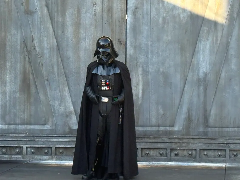 A person in a glossy black helmet, black cape and chest panel of blinking lights stands in front of tall grey metal doors.