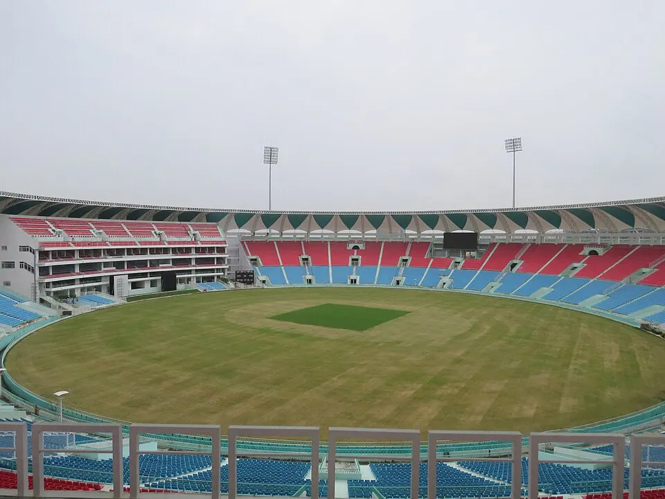 A large empty cricket stadium with red and blue seats and a green pitch strip on a brown outfield.