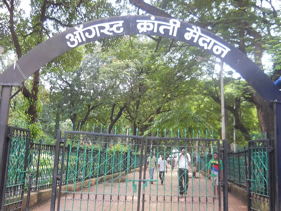 A green iron gate under an arched sign in Devanagari script, leading into a tree-shaded park path.