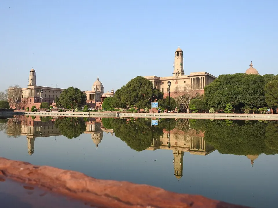 Pink sandstone government buildings with domes and towers reflected in a still pool, with green trees in between.